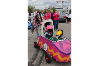 A student team pushes a handmade electric race car at the GEN‑EV competition as a helmeted driver sits inside, surrounded by other students, adults and spectators in a parking lot racecourse.