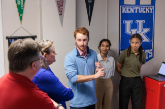 A small group stands in a campus room as a student presenter gestures while explaining a project, with University of Kentucky banners and displays visible in the background.
