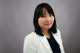 A professional portrait of a woman with shoulder-length dark hair wearing a white blazer and dark blouse, looking toward the camera against a gray background.