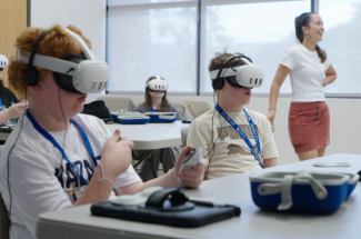 Students wearing virtual reality headsets sit at classroom tables using handheld controllers while an instructor smiles nearby. The group participates in an immersive learning experience using VR technology.