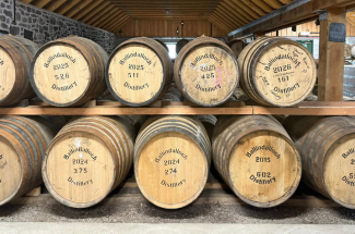 Rows of large wooden whisky barrels are stacked on wooden racks inside a distillery aging warehouse with exposed timber beams and stone walls. The barrel ends are labeled “Ballindalloch Distillery.”