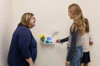 A physical therapist smiles as a young participant uses a wand to interact with a wall-mounted game station while another child looks on.