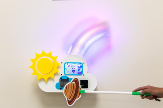A hand holds a planet-themed wand in front of a colorful wall-mounted game station featuring a lighted rainbow and animated character.