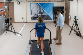 Person walks on a treadmill in a biomechanics lab while another person observes. Multiple motion-capture cameras surround the treadmill.