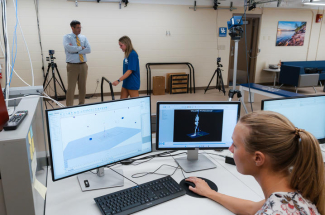 Person works at a computer displaying 3D motion-capture data while, in the background, another person walks through a biomechanics lab as an observer watches.