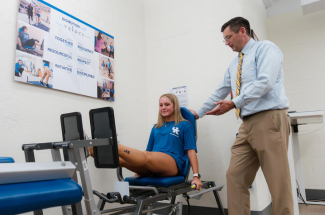 Person walks on a treadmill in a biomechanics lab while another person observes. Multiple motion-capture cameras surround the treadmill.