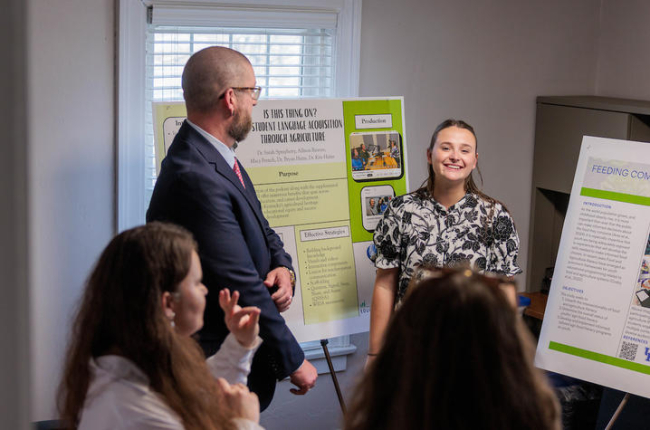 A presenter stands beside research posters and speaks to a small group gathered in a room during a poster presentation session.