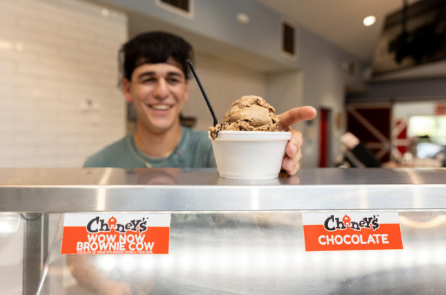 A person stands behind an ice cream display case, holding out a cup filled with two scoops of chocolate ice cream and a spoon. Flavor labels on the case read “Chaney’s Wow Now Brownie Cow” and “Chaney’s Chocolate.” 