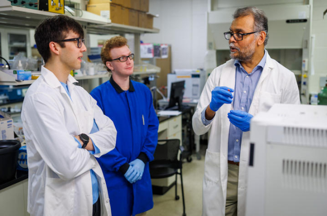 Three individuals wearing laboratory coats and protective gloves stand in a research laboratory beside benches and scientific equipment. One individual holds a small vial or tube while speaking to the other two, who are listening nearby.